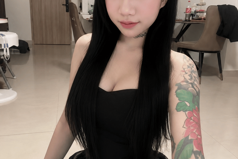 Young woman with long black hair and a floral arm tattoo wearing a black dress in a minimalist room.