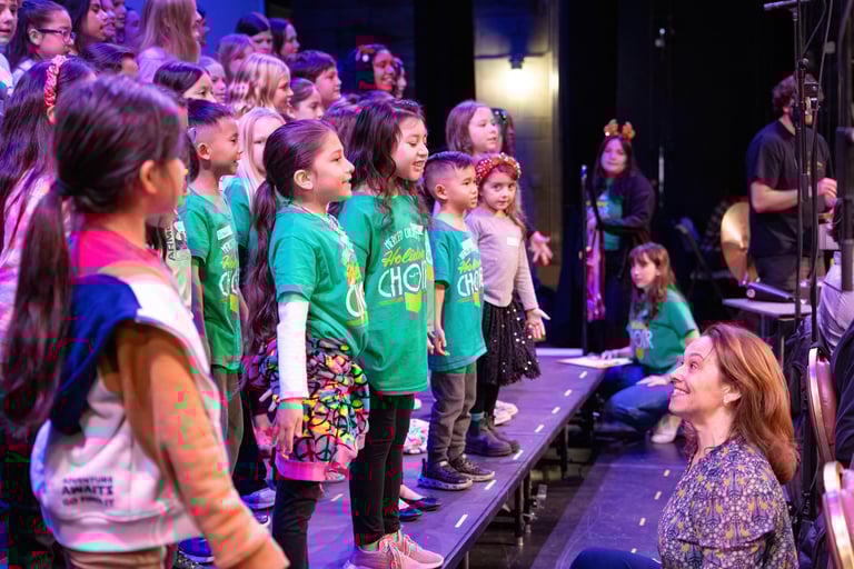 Elementary school children in green holiday choir shirts singing on stage during a winter concert.