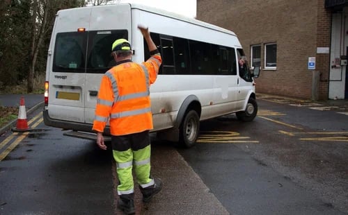 A traffic marshal in hi-vis clothing directing a white Ford Transit minibus at a commercial facility.