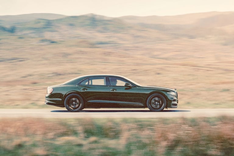 A dark green luxury Bentley Flying Spur sedan driving fast on a scenic mountain road.