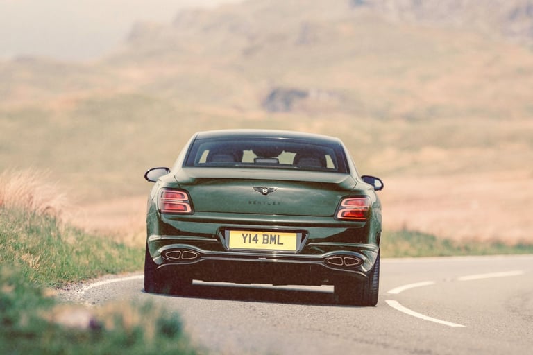 Rear view of a dark green Bentley luxury sedan driving on a winding mountain road.