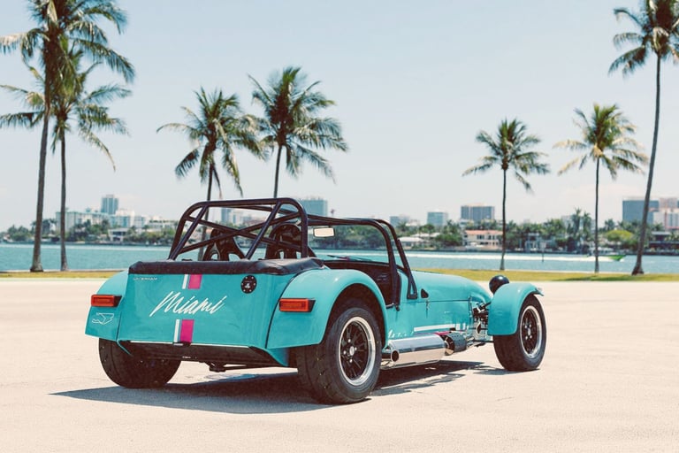 A light blue Caterham sports car parked on a Miami waterfront with tropical palm trees.