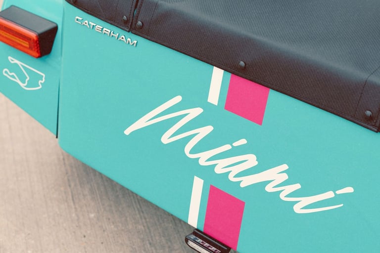 Close-up of a teal Caterham sports car featuring a Miami livery with pink and white stripes.