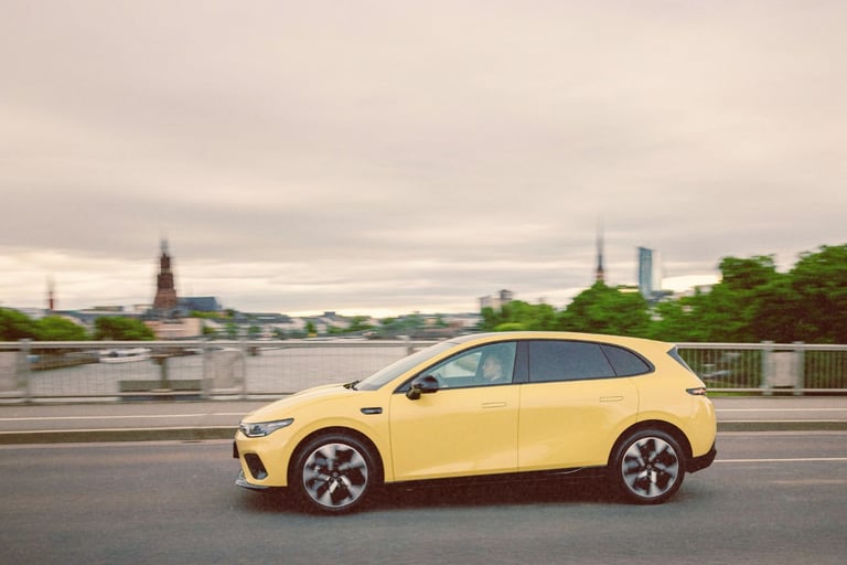 A modern yellow electric hatchback car driving across a city bridge at dusk with a blurred skyline background.