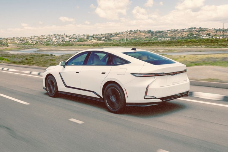White Lexus ES sedan driving on a coastal road with hills and houses in the background.