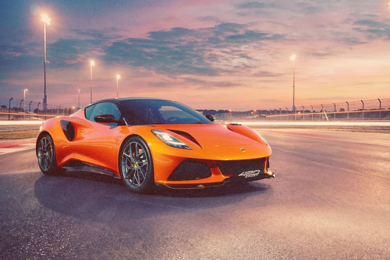 Orange Lotus Emira sports car parked on a race track at sunset with glowing stadium lights.