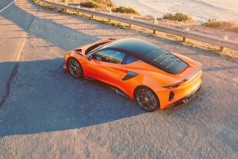 Orange Lotus Emira sports car parked on a scenic coastal road at sunset.