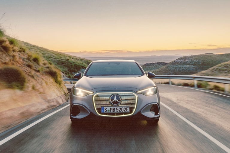 A silver Mercedes-Benz electric car driving on a winding coastal road at sunset with glowing LED grille.