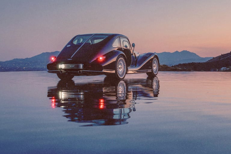 A vintage black luxury coupe reflected in water during a mountain sunset.