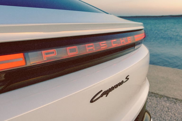 Close-up of the rear taillight and logo of a white Porsche Cayenne S parked by the ocean.