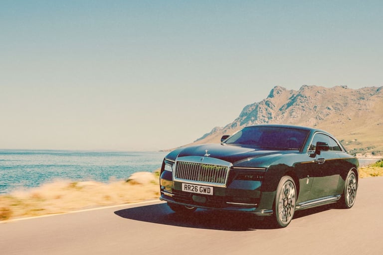 A black Rolls-Royce Spectre luxury electric car driving on a coastal road with mountains.