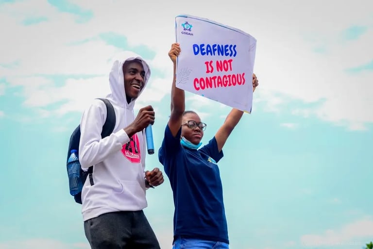 A man in a white hoodie, standing next to a woman who is holding up a banner that says "Deafness is not contagious"