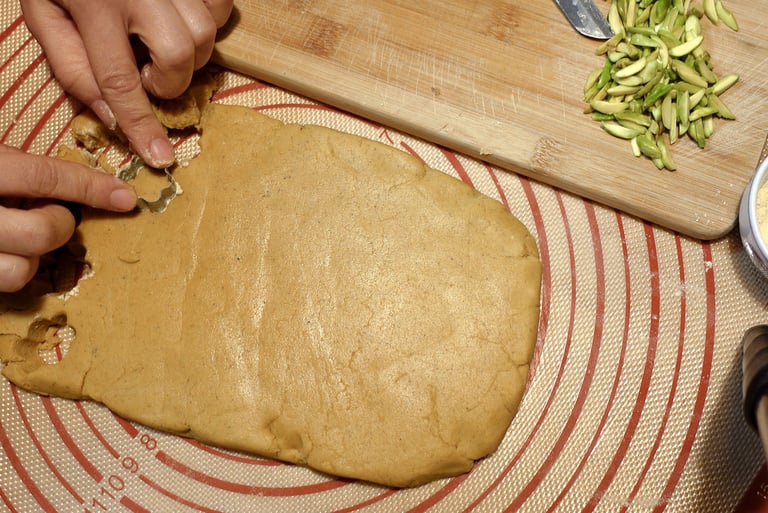 Hands cutting shapes from rolled cookie dough on a silicone mat with pistachios nearby.
