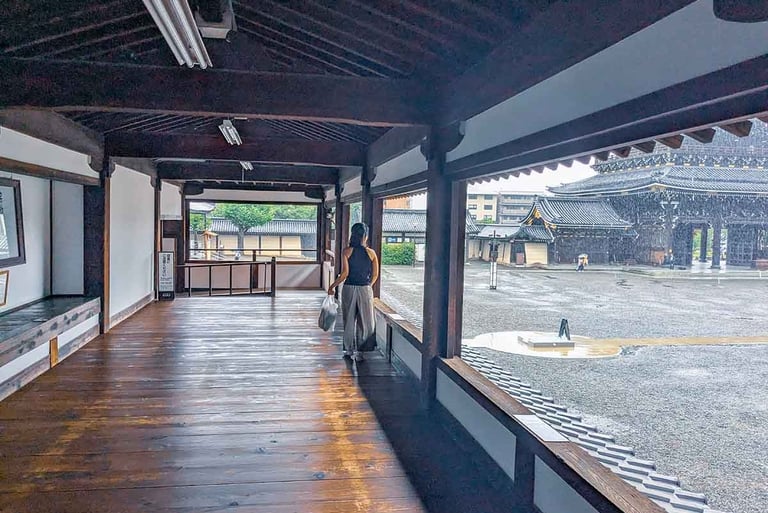 Woman walking through a covered corridor at Nishi Hongan-ji Temple while rain falls over the temple grounds in Kyoto, Japan.