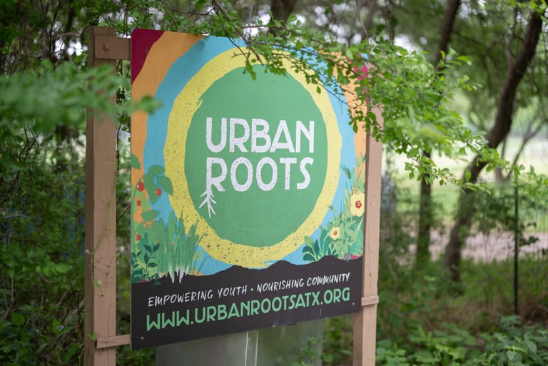 A colorful sign for Urban Roots stands nestled among lush greenery, partially framed by branches