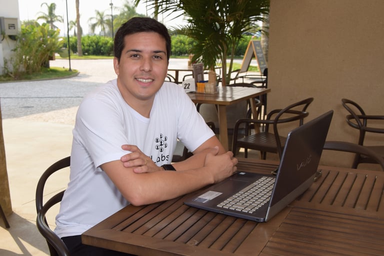 Douglas da Parceria Labs, sorrindo, sentado com notebook sob uma mesa de madeira em um café
