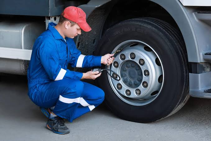 A mechanic in blue coveralls checks the tire pressure of a semi-truck wheel using a gauge.