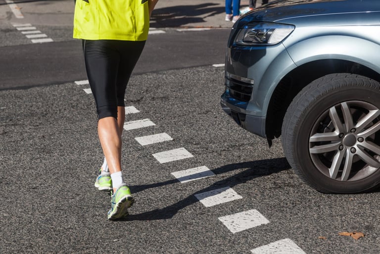 A runner in a neon yellow vest and leggings crossing a street in front of a blue car at a crosswalk.