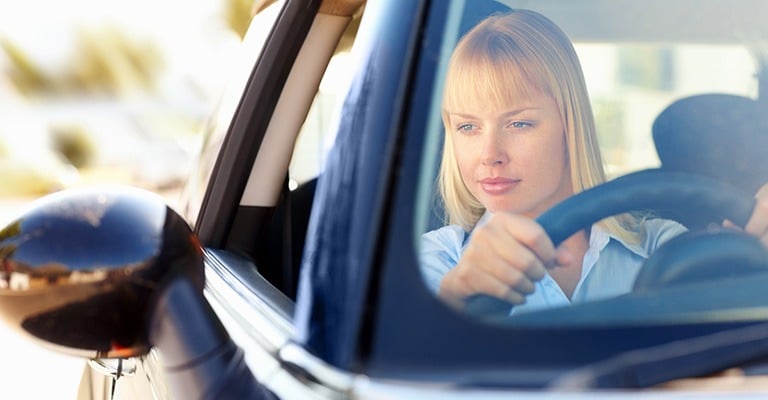 A blonde woman driving a car and looking ahead while holding the steering wheel on a sunny day.