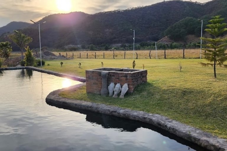 Sunset over a tranquil mountain pond with a stone barbecue pit on a lush green lawn.