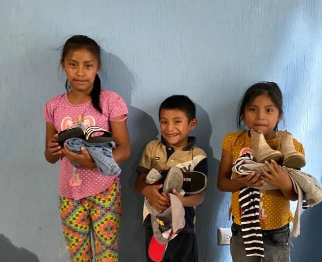 Three young children smiling while holding donated shoes and clothes from a charity donation center.
