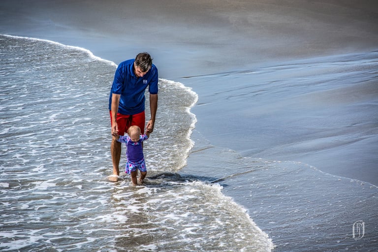 Photo of man holding up baby as they walk through water at the shore