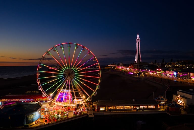 Illuminated Ferris wheel on Blackpool Central Pier at night with the Blackpool Tower in the background.