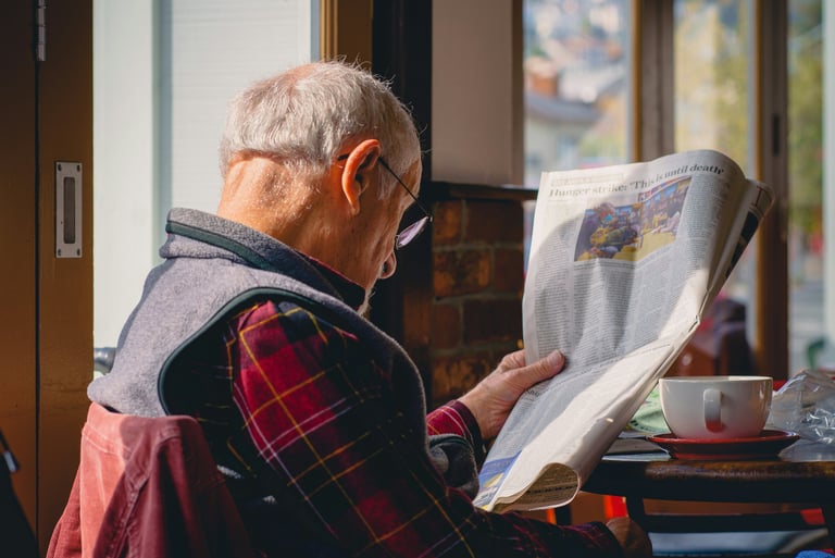 An elderly man in a plaid shirt reading a newspaper at home