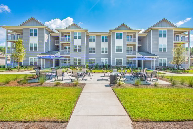 Ground-level view of outdoor patio seating with umbrellas between apartment buildings at Arrogate Village in Summerville, SC