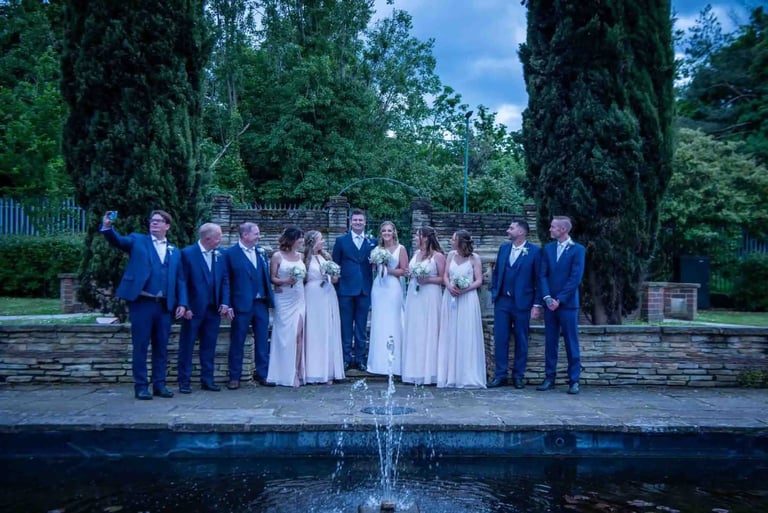 A wedding party in blue suits and pink bridesmaid dresses posing by an outdoor stone fountain.