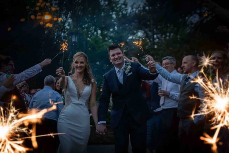 A happy bride and groom celebrate with a wedding sparkler send-off surrounded by guests outdoors at night.