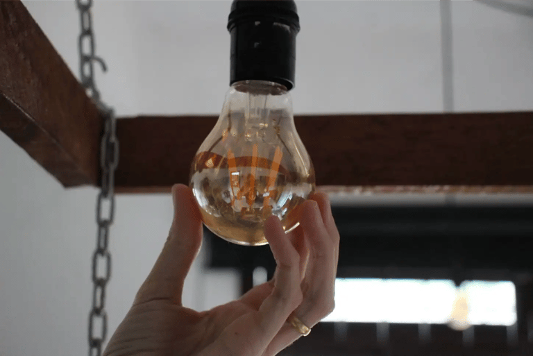 Hand holding a glowing vintage Edison light bulb hanging from a rustic wooden beam fixture.