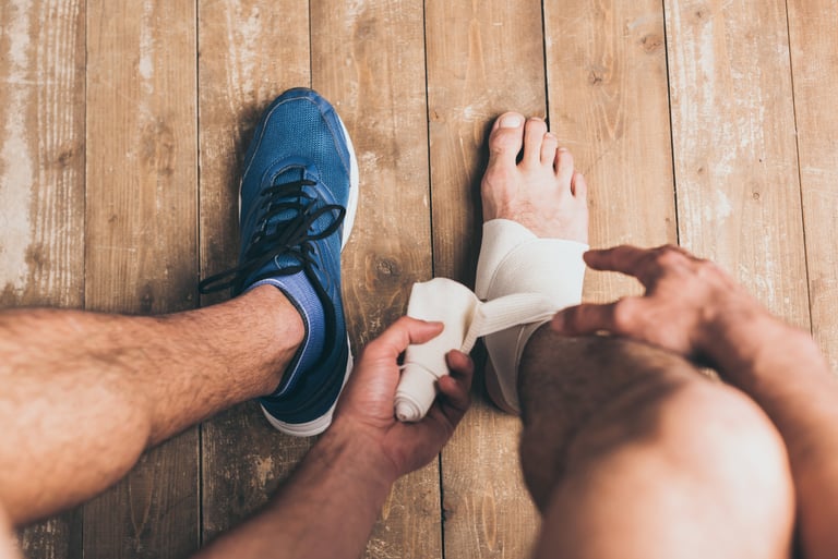 A man applying a white compression bandage to his sprained ankle on a wooden floor.