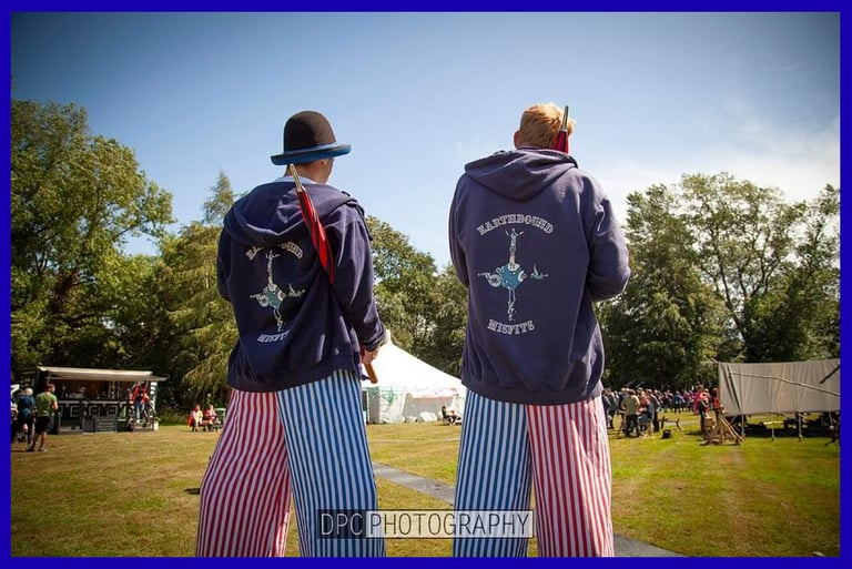 Two festival performers in striped trousers and navy hoodies stand outdoors at a summer event.