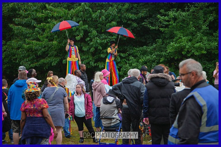 Stilt walkers in rainbow costumes holding umbrellas perform for a crowd at an outdoor community festival.