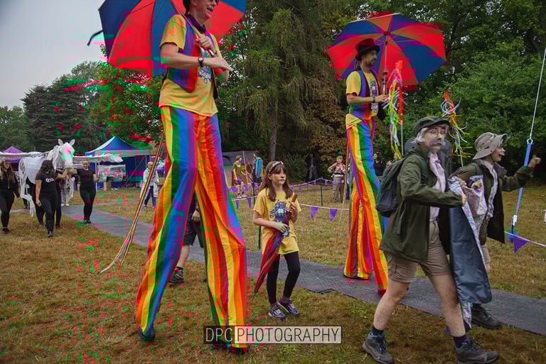 Performers on stilts wearing rainbow trousers and holding umbrellas lead a parade at a festival.