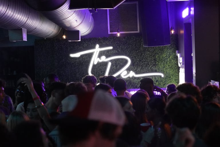 Crowded nightclub dance floor with a neon 'The Den' sign on a green foliage wall.
