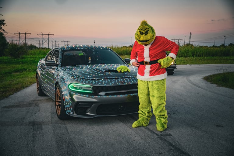 A person in a Grinch costume posing next to a Dodge Charger wrapped in Christmas lights at sunset.