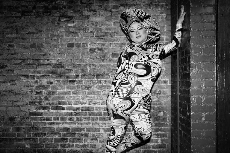black & white drag queen posed fashionably in front of brick wall on stage in a patterned bodysuit