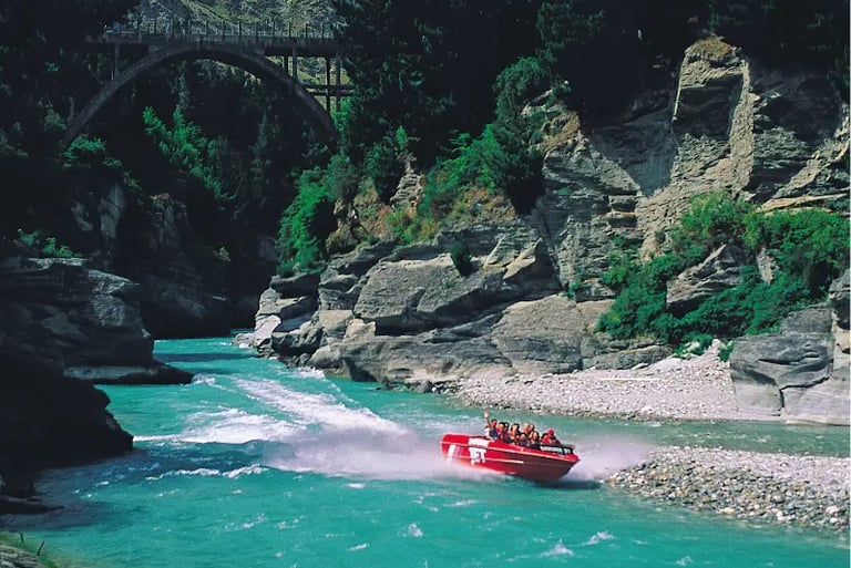 A red Shotover Jet boat speeding through the turquoise Shotover River canyon in Queenstown, New Zealand.