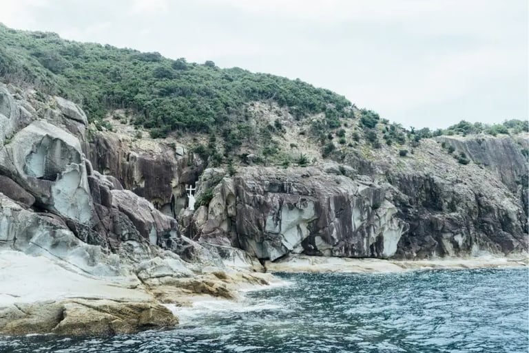 A white religious statue stands on a rocky cliffside along a rugged coastline by the sea.