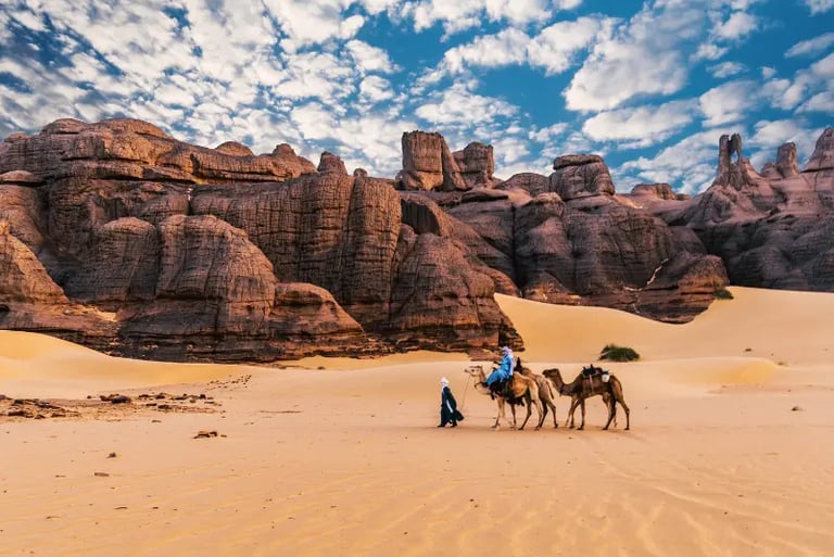 A camel caravan guided by people through the Sahara Desert sand dunes with rocky mountains.