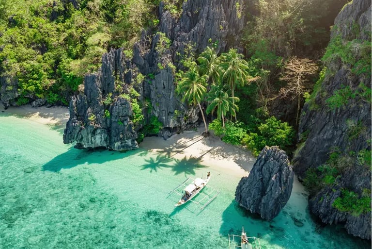 Aerial view of a tropical El Nido beach with limestone cliffs, palm trees, and traditional boats in clear water.