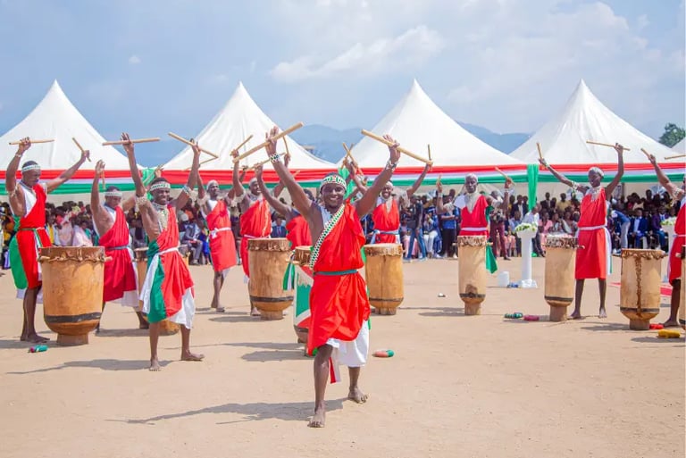 Burundi drummers in traditional red and white robes perform a rhythmic dance with wooden sticks.