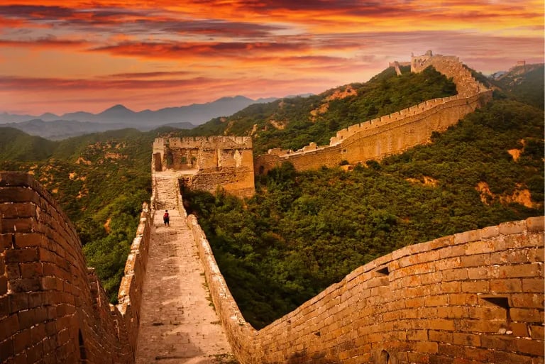 The Great Wall of China winding through green mountains under a vibrant orange sunset sky.