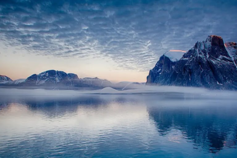 Dramatic Arctic landscape featuring snow-capped mountains reflecting in calm blue water under a textured sky.