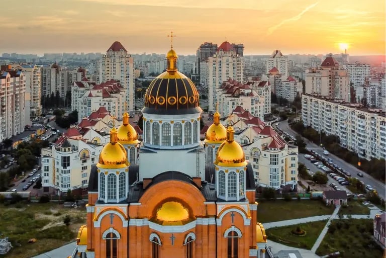 Golden-domed Orthodox cathedral overlooking the Kyiv city skyline and residential buildings at sunset.