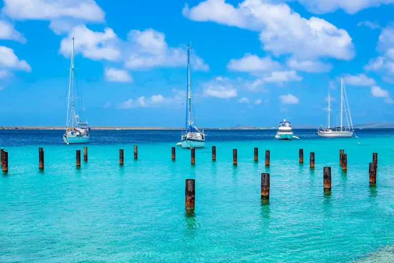 White sailboats anchored in tropical turquoise water with wooden pier pilings and a blue sky.
