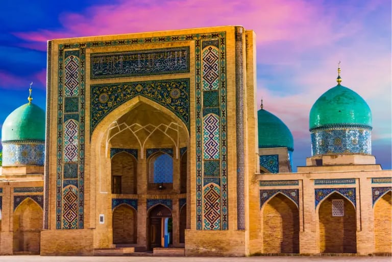The historic Hast Imam Ensemble in Tashkent, Uzbekistan, featuring blue tiled domes and mosaic arches at sunset.