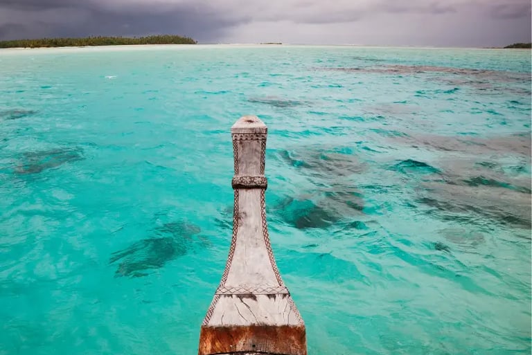 Traditional wooden boat bow sailing through clear turquoise tropical ocean waters under a cloudy sky.